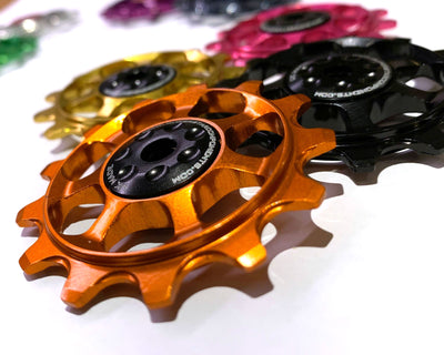 A set of high-performance golden jockey wheels with a black center, designed for a 12-speed Shimano rear derailleur system, displayed alongside other colored jockey wheels in the background.