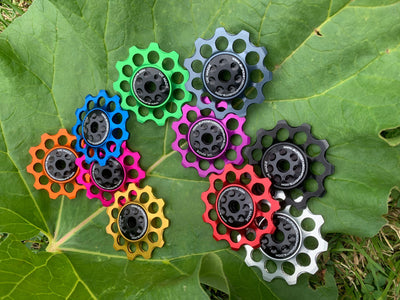 A collection of colorful jockey wheels for bicycles, displayed on a green leaf. The wheels are available in various colors including black, blue, green, red, and yellow.